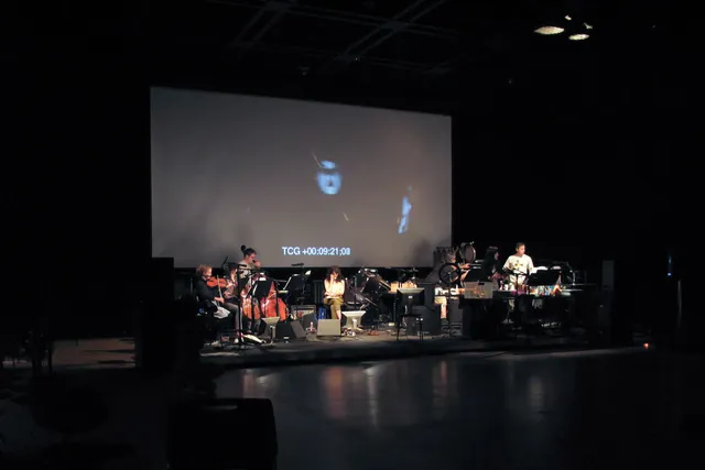 A small pit orchestra set up on a black stage un a large screen projecting "TCG +00:09:21;08" in white block letters.  