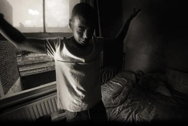 A young Black man standing in an apartment bedroom next to an unmade bed with arms outstretched to the sides. 