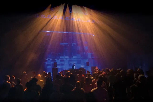 A man DJing on a small stage lit my blue and yellow lights in front of a silhouetted crowd. 