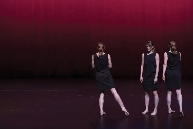 Three female dancers wearing black one shoulder dresses in a tight formation on a stage lit softly in burgundy light. 