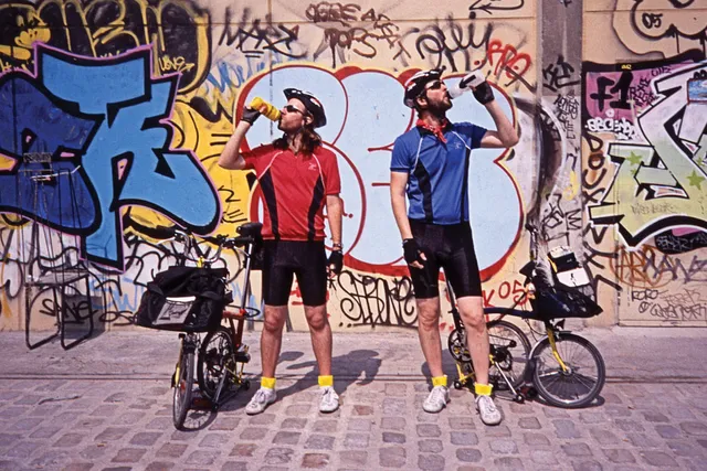 Two male bicyclists, one wearing red the other blue drinking from their water bottles with a wall of graffiti in the background. 