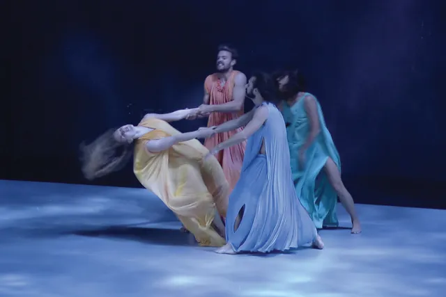 Four dancers wearing pastel colored flowing costumes standing in a circle. A woman in yellow throws herself back dramatically as the others hold her arms. 