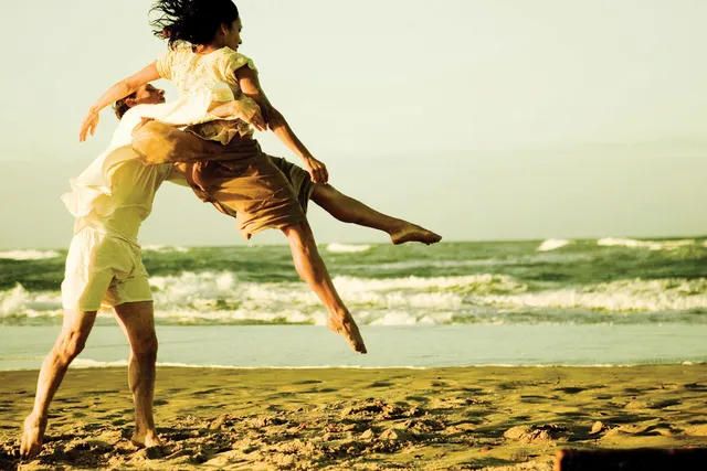 A man dressed casually tosses a woman into the air on a beach lit by warm yellow light. 