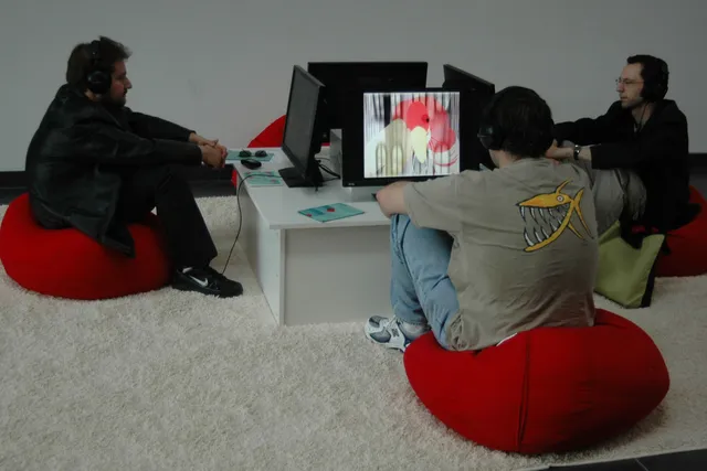 Three men seated on small red bean bag chairs on a white shag rug watching individual screens showing abstract images. 