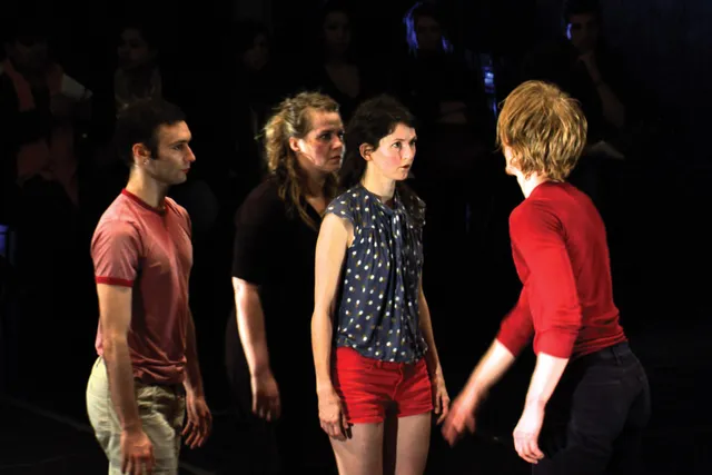 Four dancers dressed in street clothes with red accents on a dark stage. One person wearing a red long sleeved shirt with the back to the viewer, reaches towards another dancer wearing red shorts as the two others look on. 