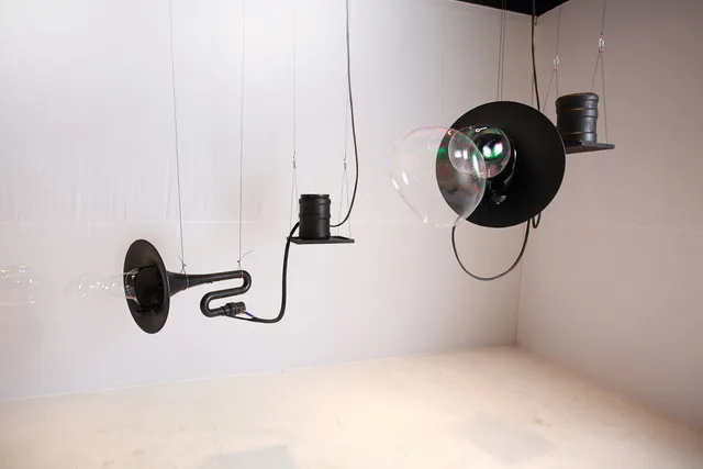 Two large black tuba-like horns blowing bubbles suspended by wire in a white gallery. 