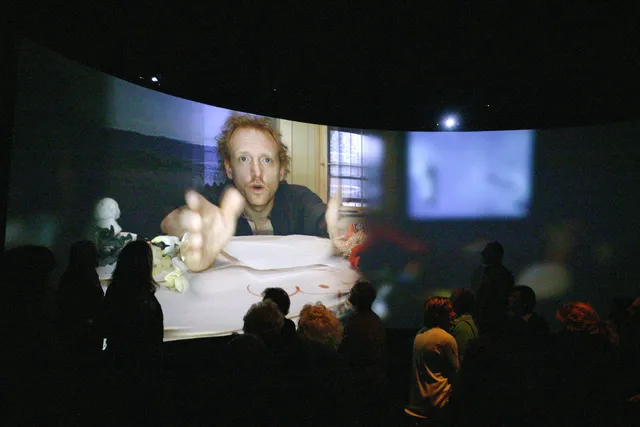 A small crowd milling about in front of a panoramic screen showing an image of a man with red hair reaching. 