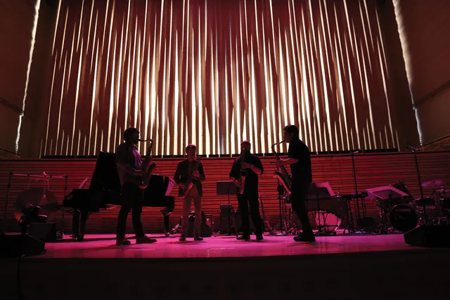 Four men playing saxophone in a semi circle on a stage washed in pink light. 