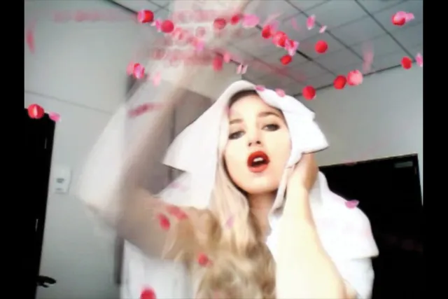 A white woman with platinum blonde hair and red lipstick throwing rose petals into the air while wearing a white piece of cloth on her head. 