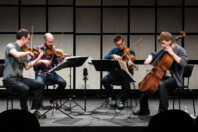A string quartet of four white men playing in front of a wall of gray acoustic tiles. 