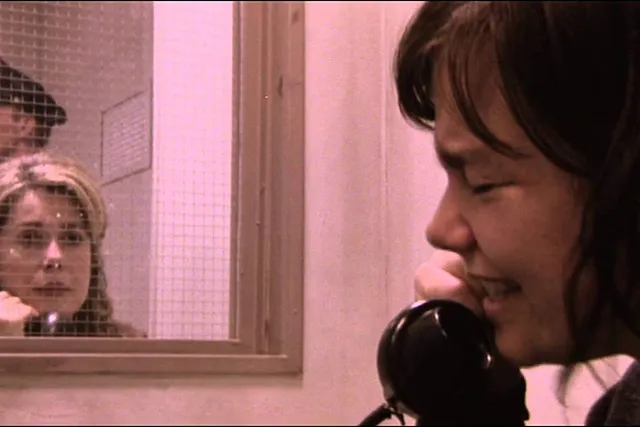 A  woman crying into the phone as another sitting behind prison glass listens. A guard stands in the background. 