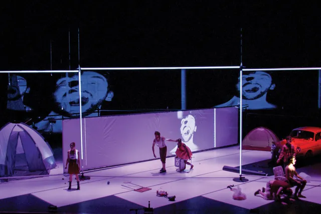 Six performers on a light purple stage with a vintage car, several tents, and other found objects.  Three performers are the front of the stage, one screaming theatrically. Two performers are behind them walking hunched towards the sixth performer.