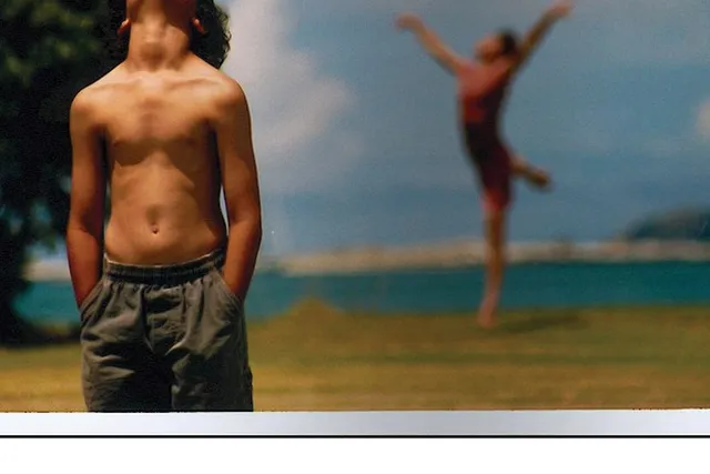 A young boy standing on a beach with tanned skin wearing a gray bathing suit looking up with his head cut out of the image. Behind him another person can be seen leaping. 