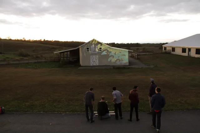 Six people standing in a field looking at a projection of black waves on a barn. 