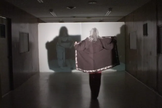 a woman in a gray room creates a shadow by holding open her jacket against a screen. 