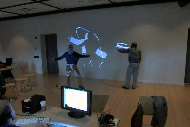 Two people with backs to the viewer interacting with a wall projection controlled by a bald man sitting at a computer in the foreground. 