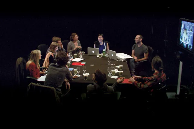 A group of nine people sitting around a conference table in active discussion in a dark room. 