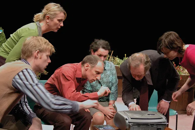 A group of six adults gather and hunched over a boombox on a dark stage with potted greenery. 