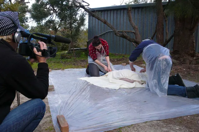 Kathy High filming a deceased pig being prepared for burial on a sheet of plastic. A man and woman, both wearing plastic aprons gently unwrap the pig from a white cloth under a tree behind a blue garage. 