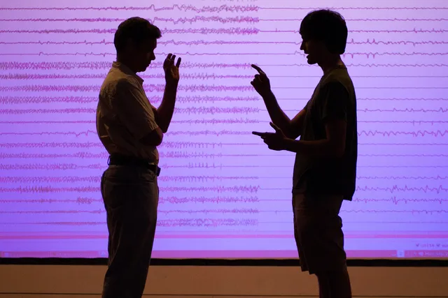 Two men standing in front of a pink projection of sound waveforms.