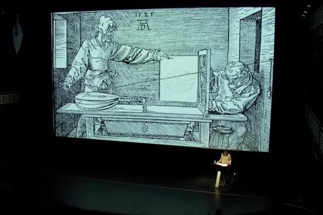 a woman giving a talk on a black stage in front of a projection of a 1500's sketch. 