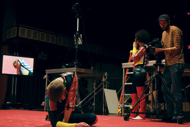 A dancer wearing a colorful costume laying on a red carpet while being filmed as Maria Hassabi looks at the video feed on a screen, contemplating. 