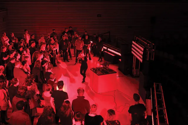 Pharmakon performing on the concert hall stage with an audience standing around her washed in red light. 