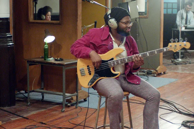 A Black man wearing a beanie and dressed in 1970's fashion plays the bass in the middle of a 70's era recoding studio. 