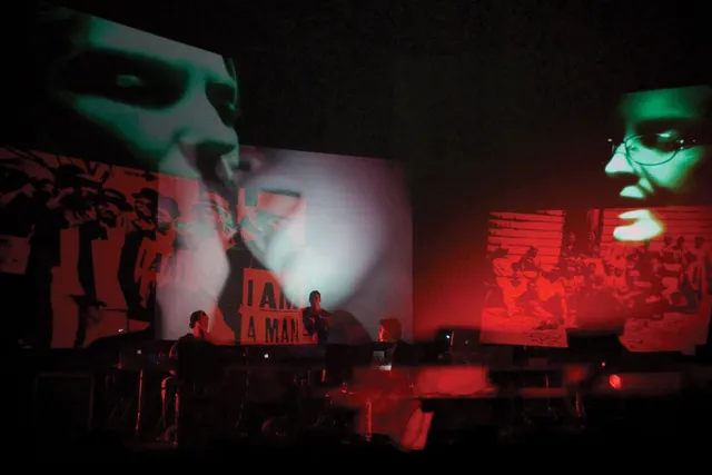 Three faces in green projected onto a dark stage with red images of protesting overlapped. A small pit orchestra is in front of them. 