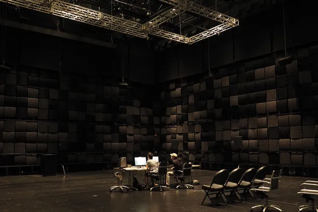 A black box studio with black acoustic tiles on the wall. Two people sit in the middle of a room at a desk with two computers below a large rectangular grid suspended from he ceiling. 