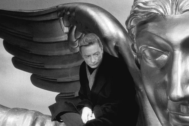 A man wearing a black overcoat looking pensively over his shoulder sitting beneath the wing of a giant metallic angel statue. 