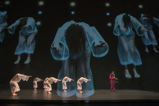 Six dancers dressed in sheer kaftans on stage with a stedicam operator wearing pink in front of a projection of previously mentioned dancers