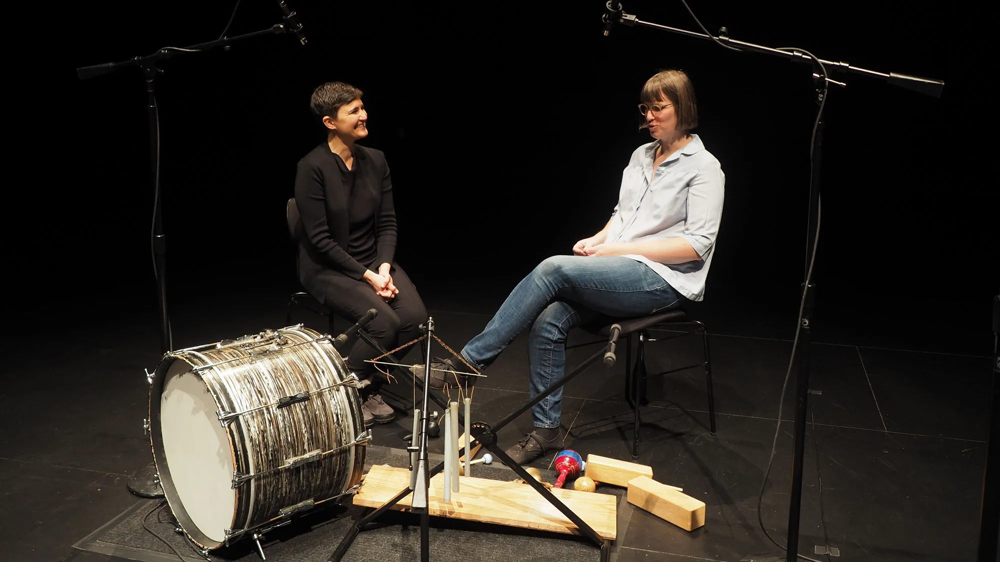 sara hennies and anne leilehua lanzilotti in conversation on a dark stage behind a kick drum. 