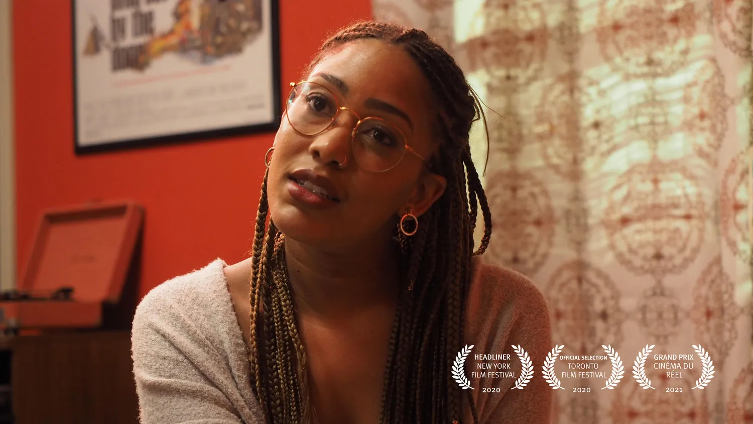 A Black woman wearing glasses and long braids looks toward the camera with head cocked in a room with red walls and patterned drapes. 