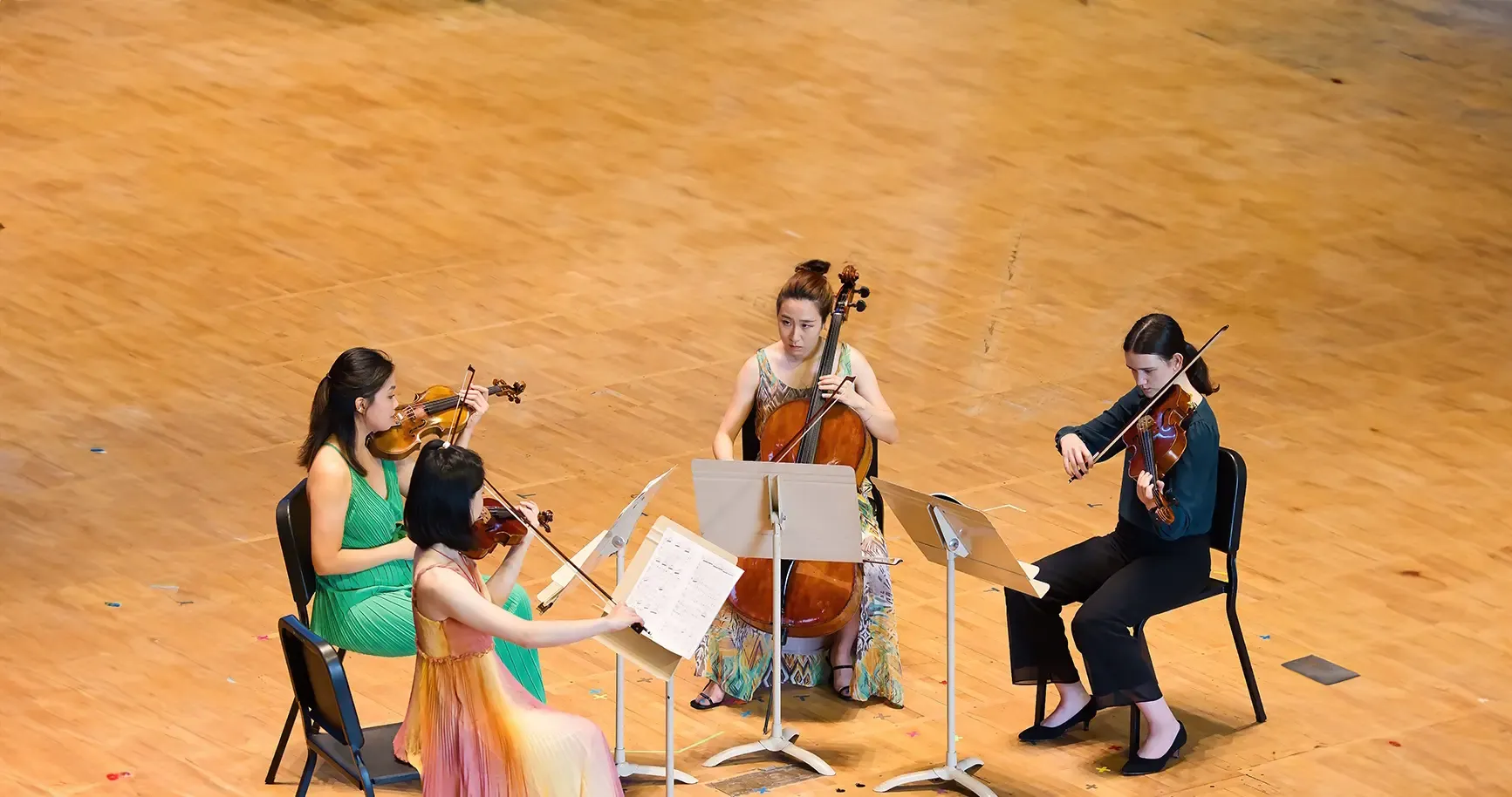 Four musicians performing in a string quartet setting on a wooden stage facing each other, two playing violins, one playing the cello, and one reading sheet music while playing the viola.