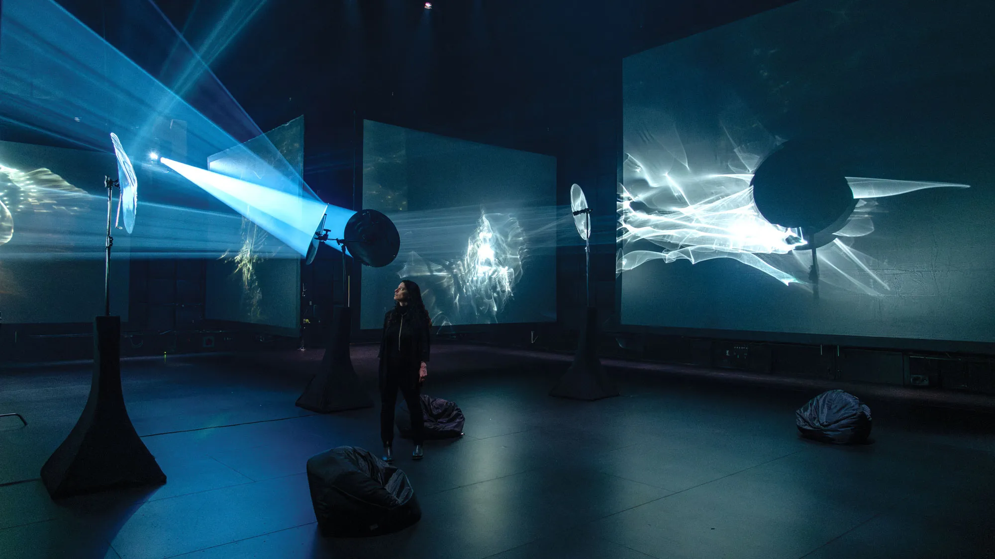 Yael Erel stands in studio 1 with light projected throughout on screens.