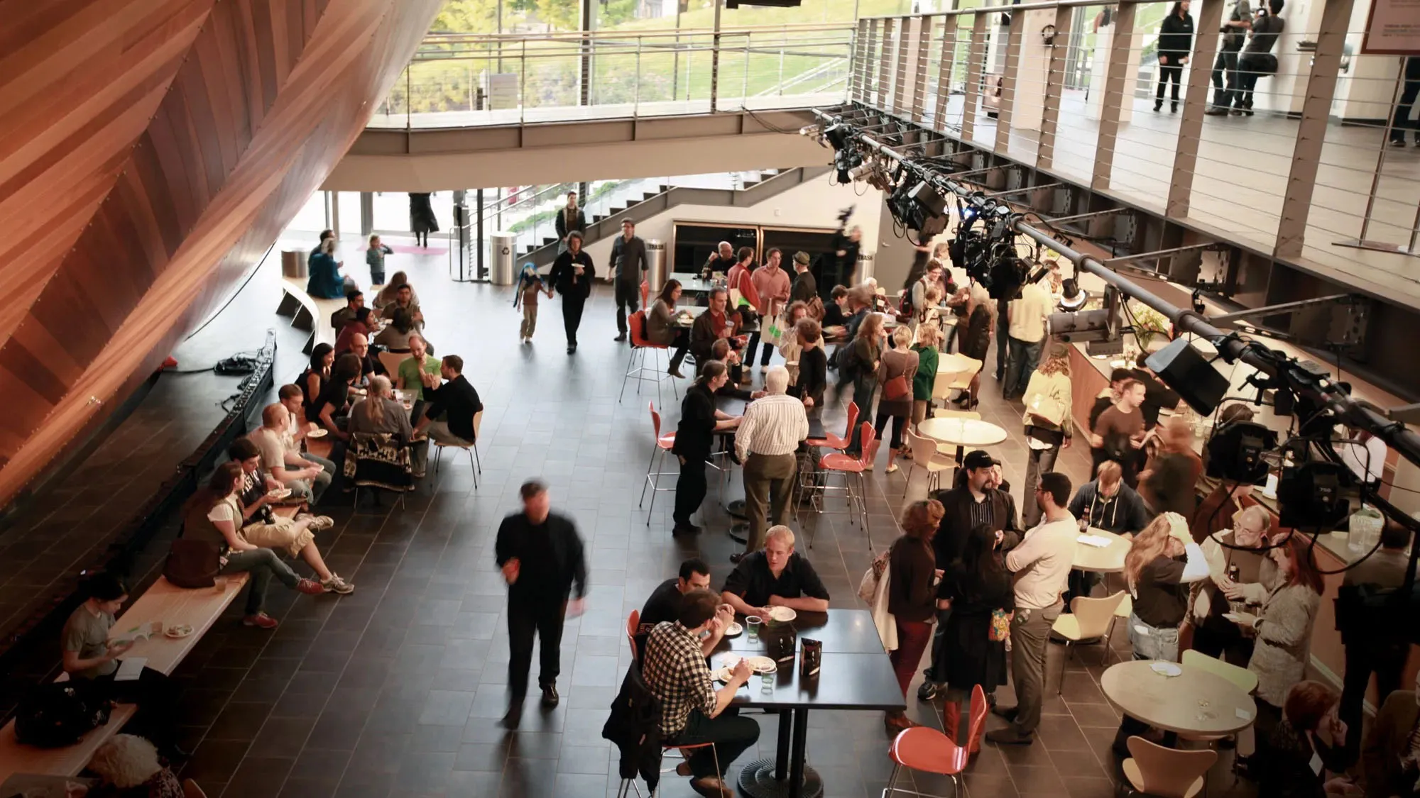A crowd of people seated on on benches, at tables, and milling about in a cafe. 