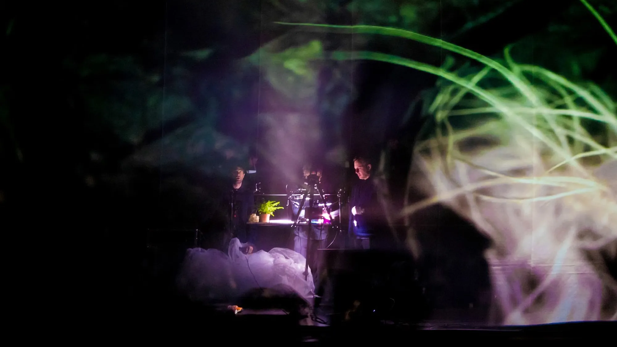 abstract shapes on a scrim in the foreground bathe two musicians sitting at a table working on their computers in the background in a black studio.