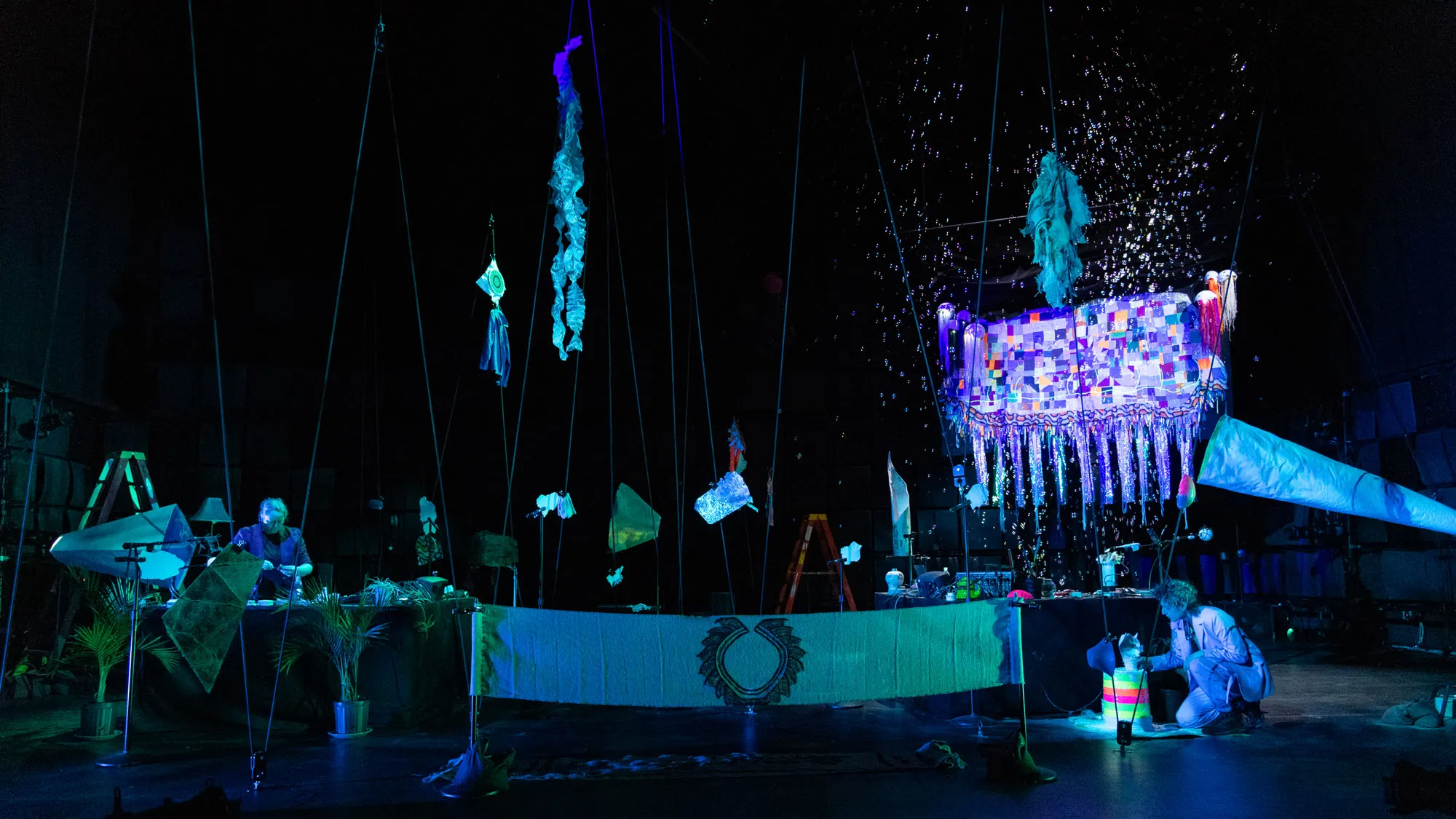 two women in a black box studio doing foley with a glow in the dark jellyfish quilt suspended high in the background with bubbles flying all over