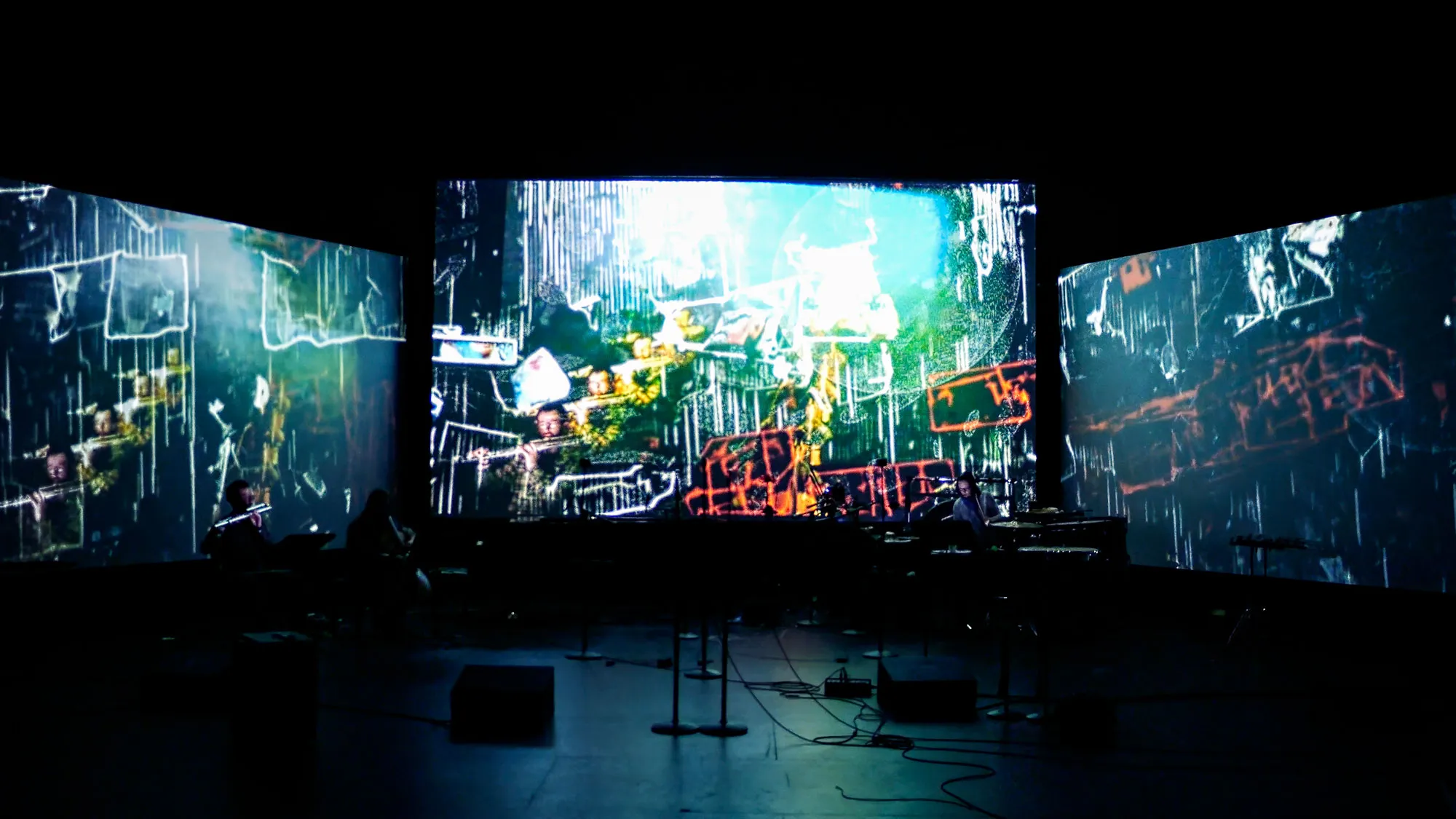 three screens with projections in the background of a band in the foreground of a black box studio.