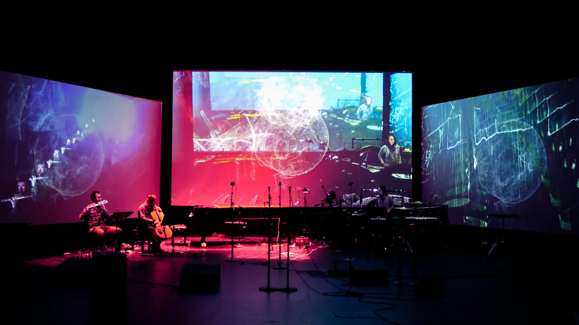 three large screens with projections in a black box studio with musicians performing. 