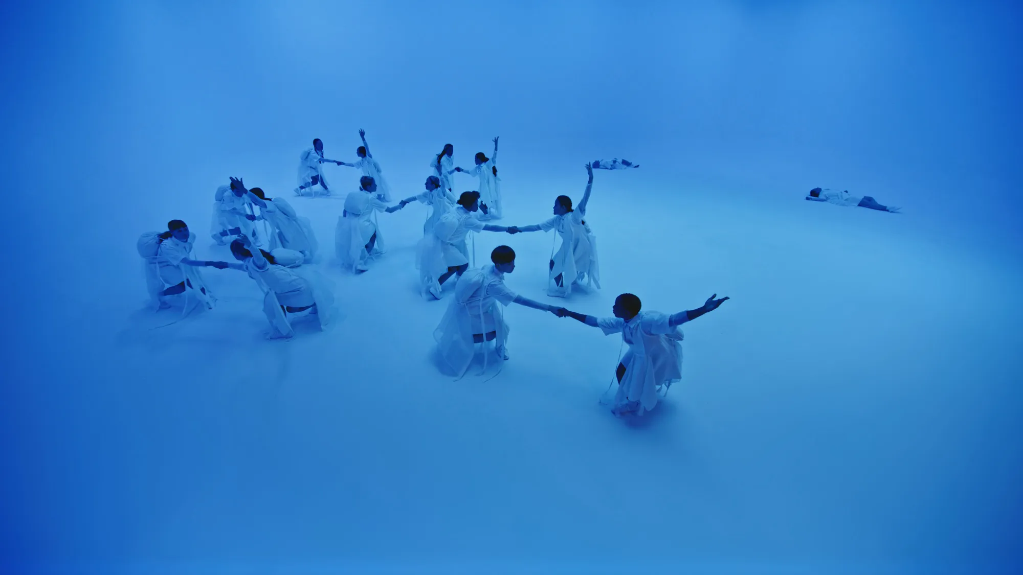 a group of dancers in white in a formation in a white cube bathed in blue light