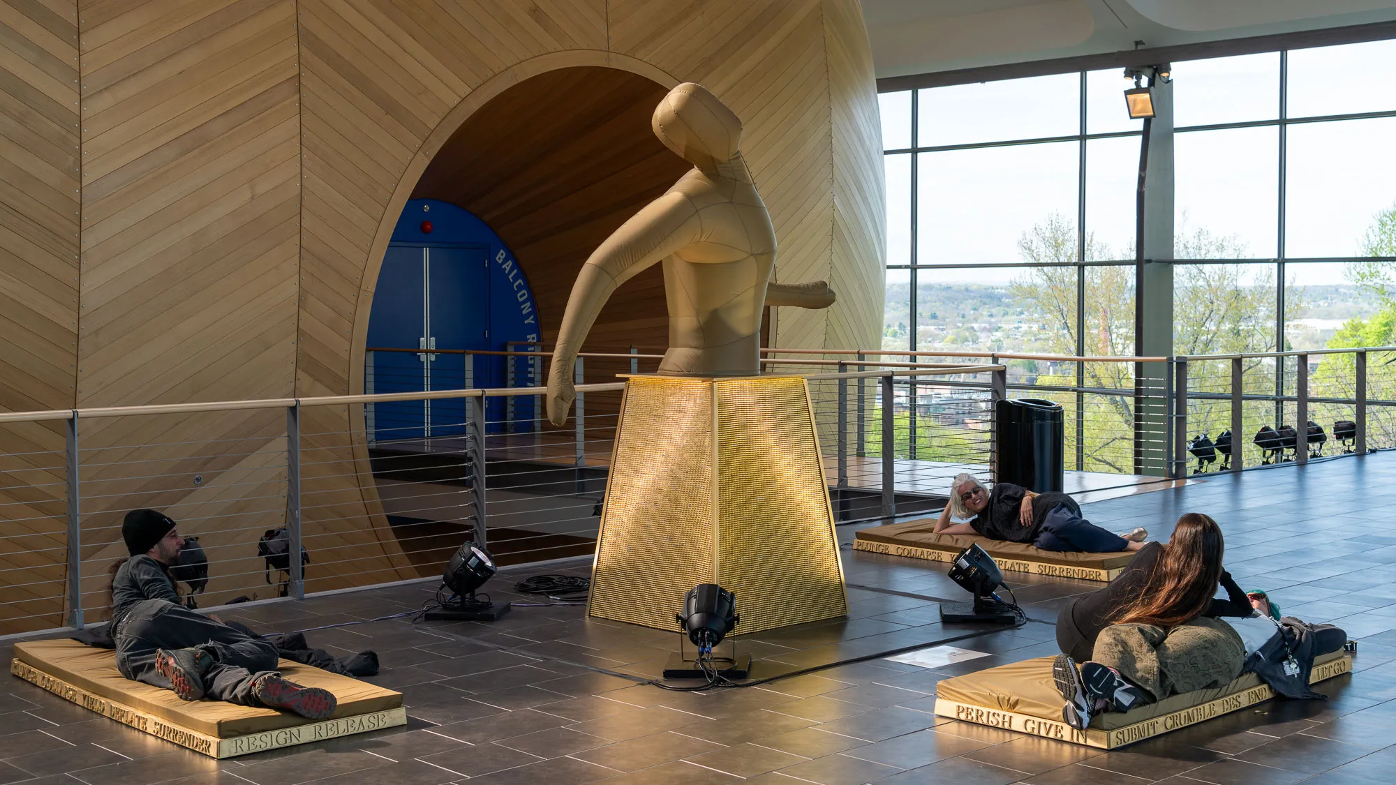 an inflatable sculpture of an amorphus human in a building lobby with people laying on platforms in the foreground