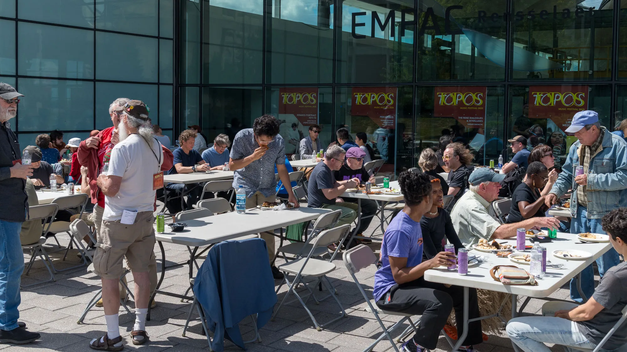 many people sitting at long tables in front of a glass building eating on a sunny day