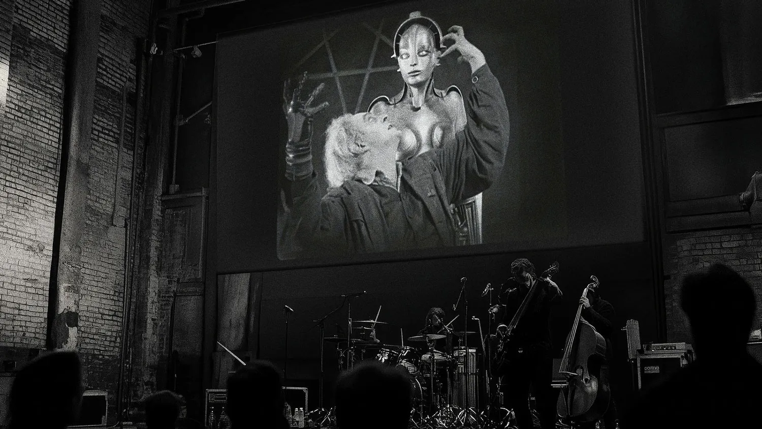 A black and white photograph of two musicians, [l-r] a guitarist and drummer, performing beneath a large screen on which a celebrated frame from Fritz Lang’s silent film Metropolis is projected.