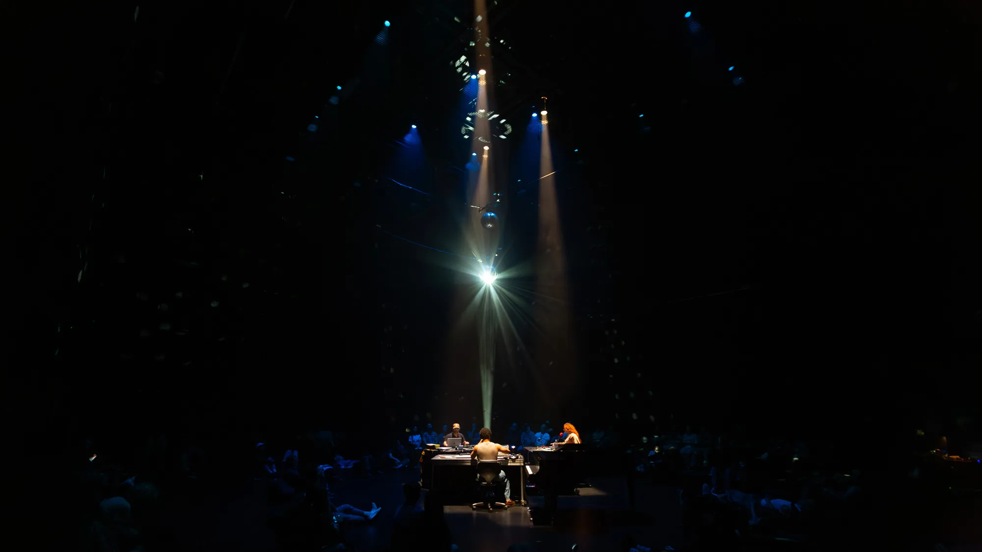 three lights show down on a disco ball as three performers in a circle lay down a DJ set in a black studio. 