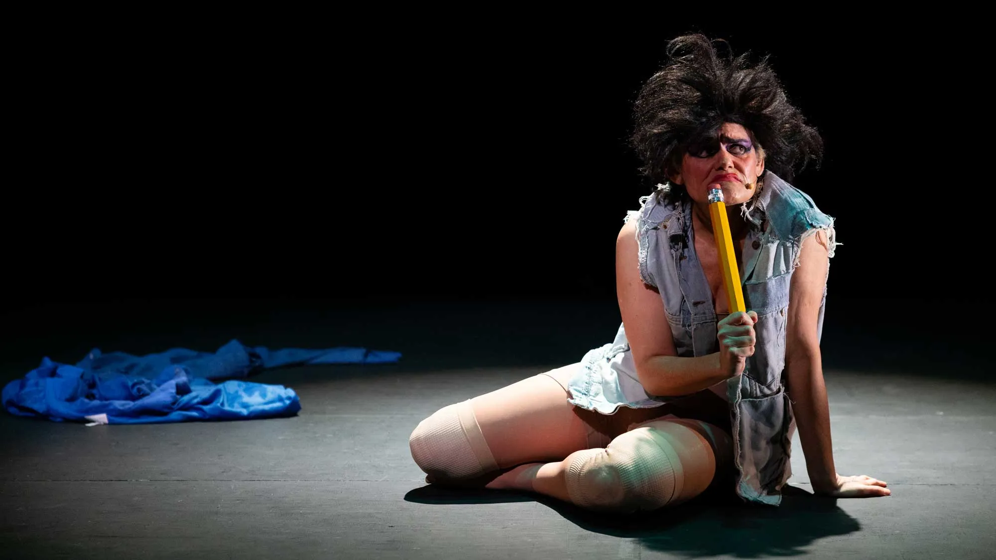 Dynasty Handbag sits on a dark stage in a torn denim vest, thigh-high stockings, and voluminous black wig, holding an oversized yellow pencil to her mouth like a microphone. A crumpled blue garment lies on the otherwise bare stage behind her.