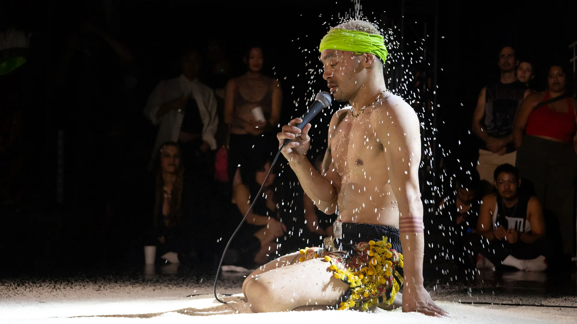 A shirtless performer kneels on a sandy stage, speaking into a microphone while a shower of sand or small particles falls around them. The audience, visible in the background, watches intently.
