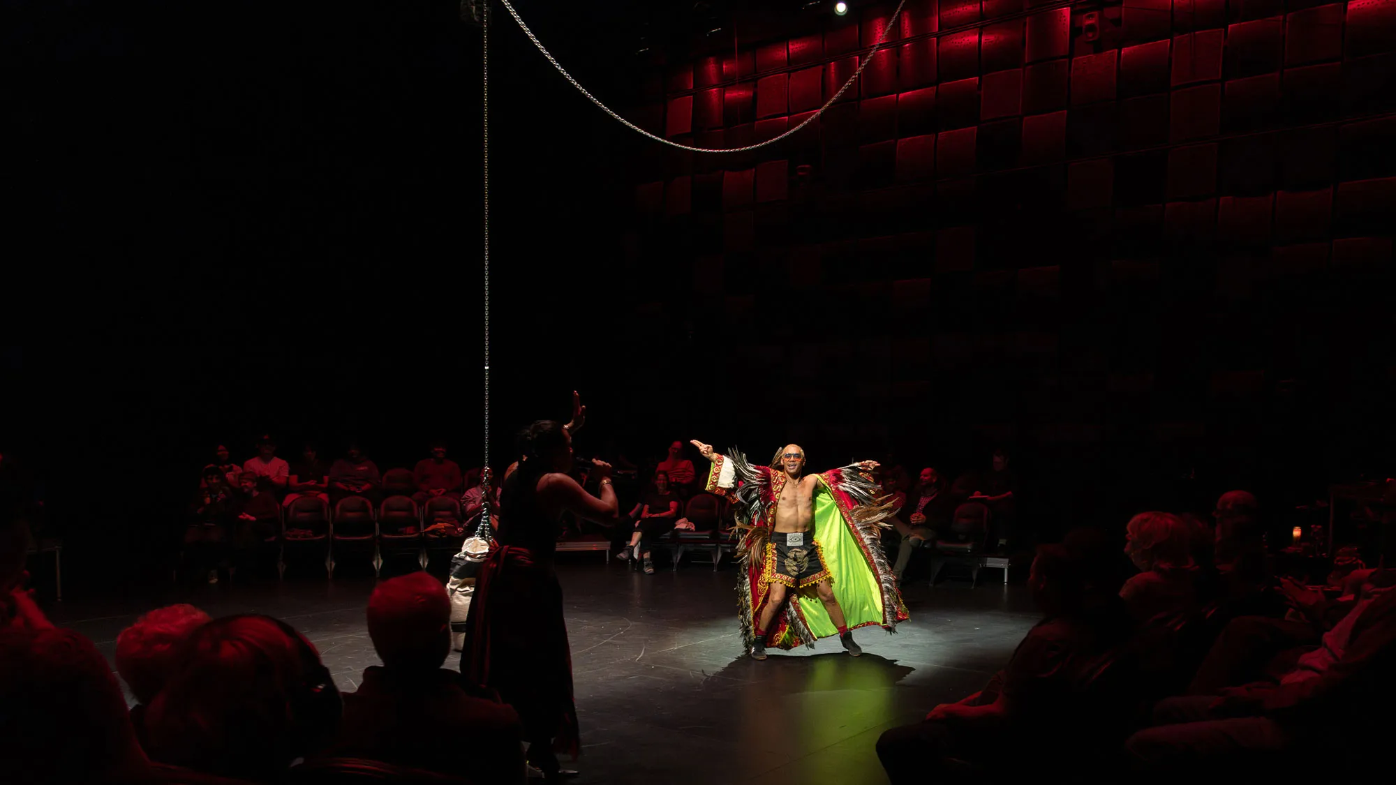 a filipino performer in a indigenous cape with chicken feathers spreads his arms wide near a hanging punching bag surrounded by audience in 360 degrees.