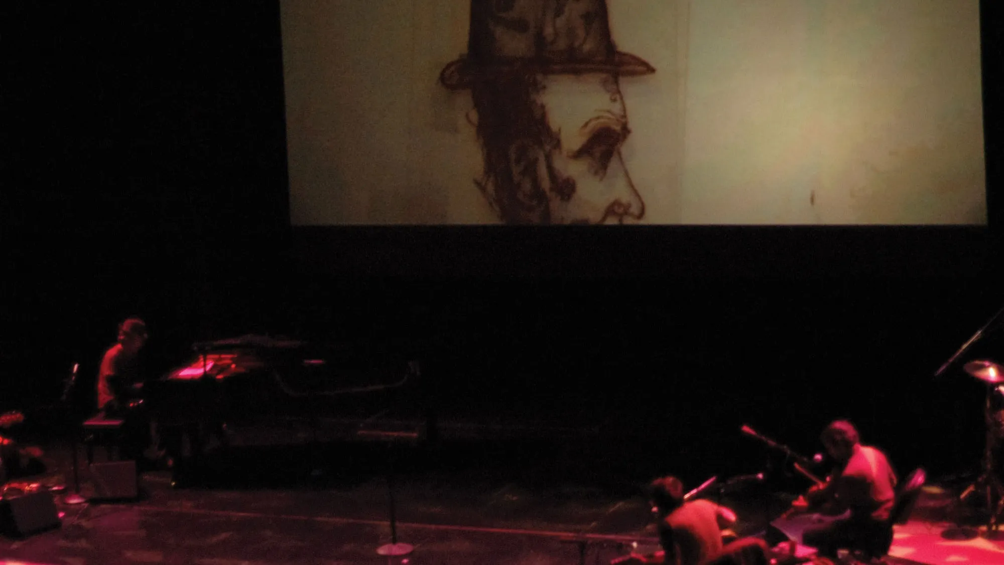 A small pit orchestra dimly lit in red and blurred in motion, playing at the base of a movie screen showing a sketch of profile of a man wearing a bowler hat.